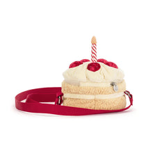 A plush cake-shaped bag with cream-colored frosting, red cherry details, a striped candle, and a red strap, shown on a plain white background. Behind view.