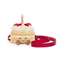 A plush cake-shaped bag with cream-colored frosting, red cherry details, a striped candle, and a red strap, shown on a plain white background. front view.