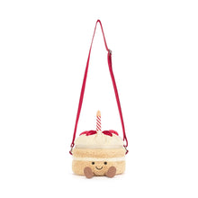 A plush cake-shaped bag with cream-colored frosting, red cherry details, a striped candle, and a red strap, shown on a plain white background. Strap extended view. 